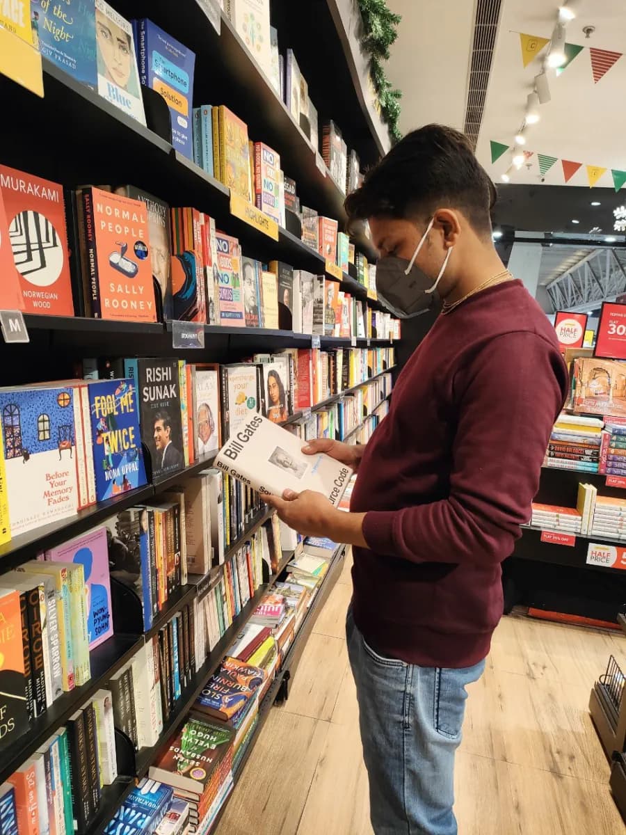 Chandan in a bookstore wearing a face mask and a maroon sweatshirt, browsing a book in front of tall shelves.