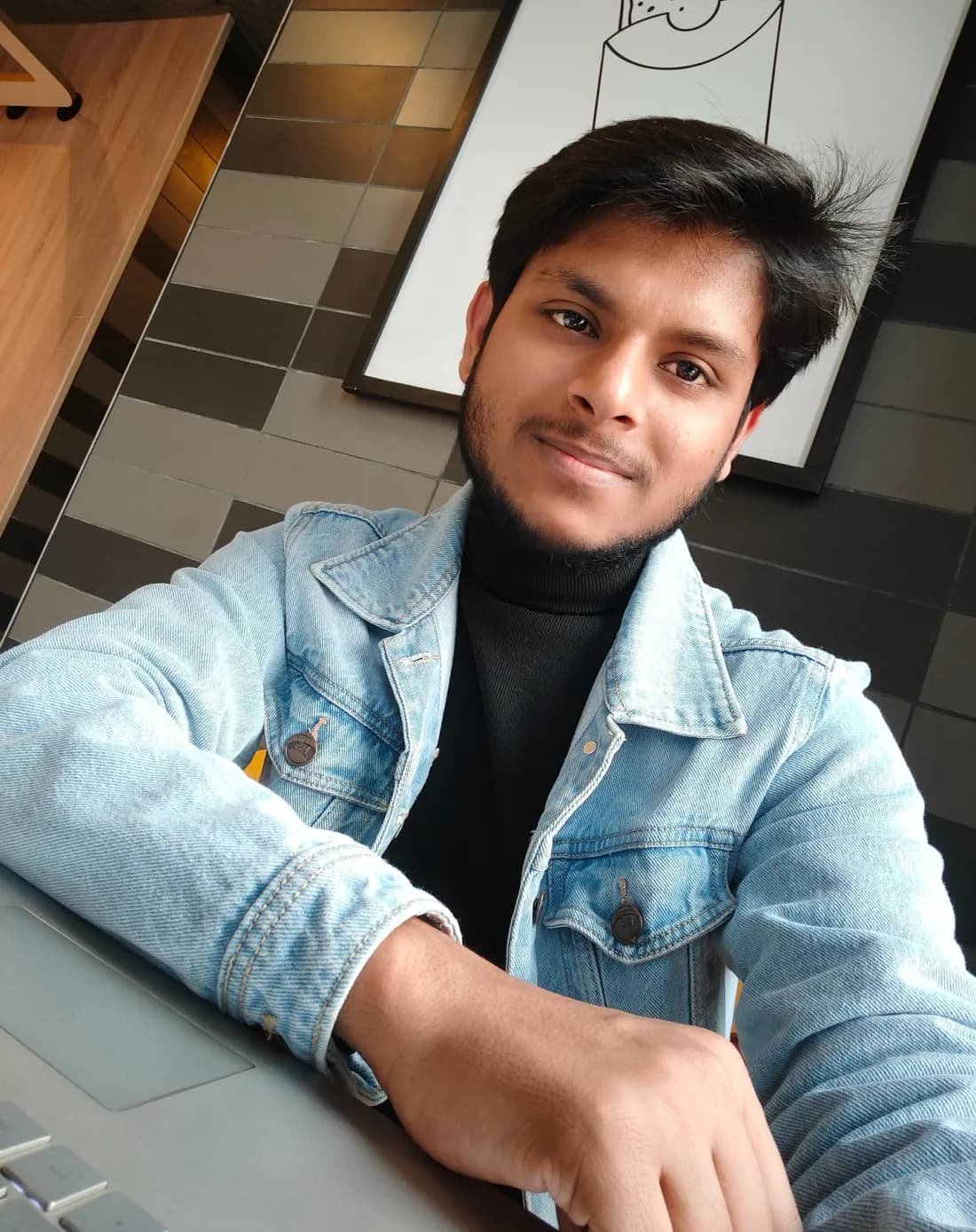 Chandan smiling at the camera in a light denim jacket and black turtleneck, with a laptop in front of him at a café.