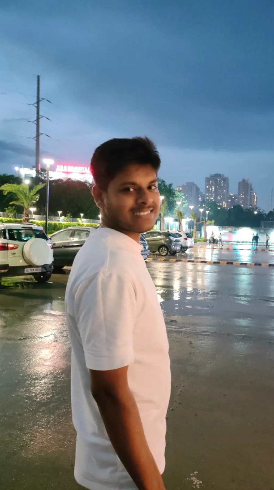 Chandan in a white t-shirt glancing back over his shoulder with a big smile, on a rain-wet street at dusk with city lights behind him.